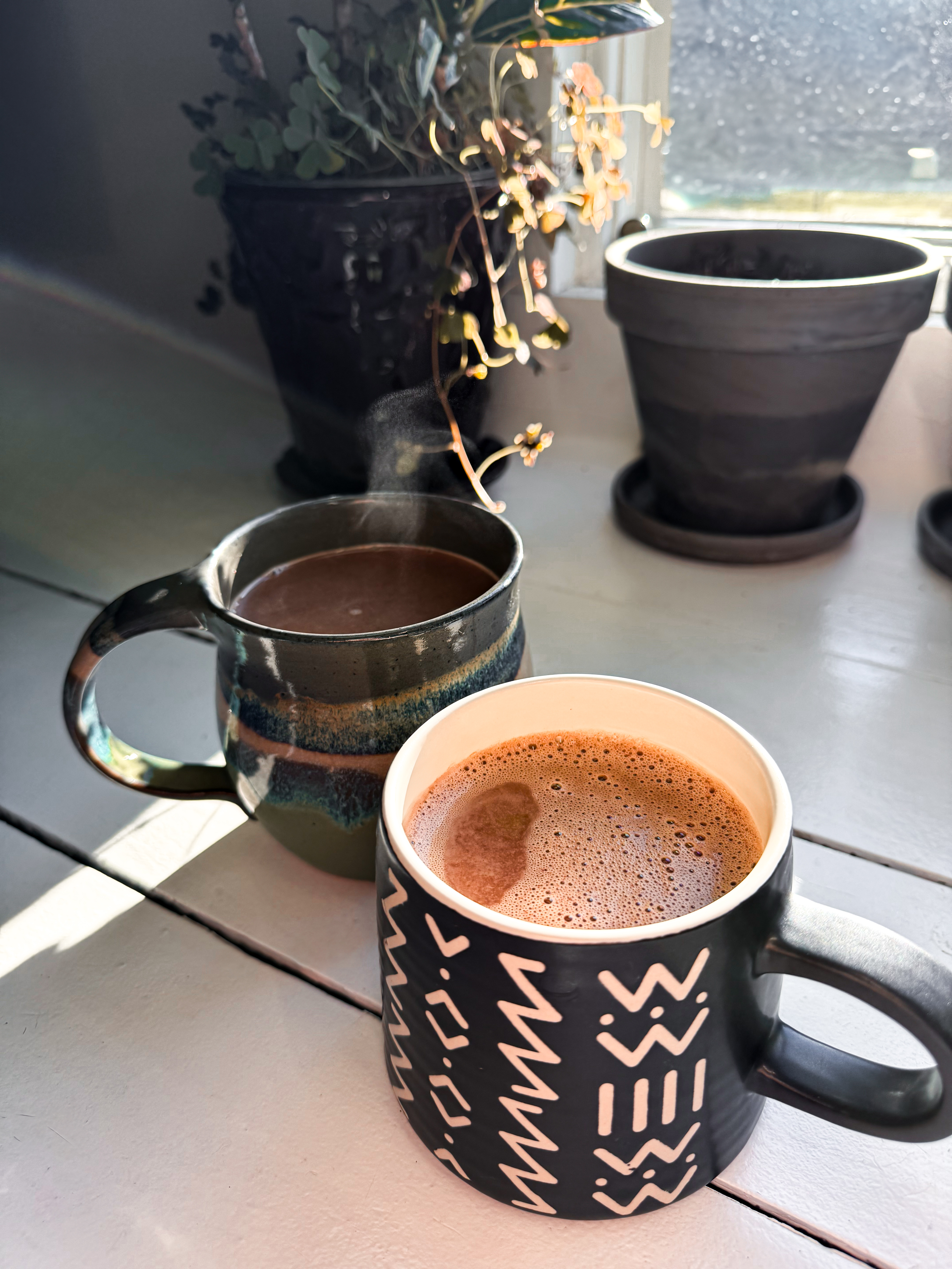 Two mugs of chicory hot chocolate on a wooden windowsill, with natural light and potted plants in the background.