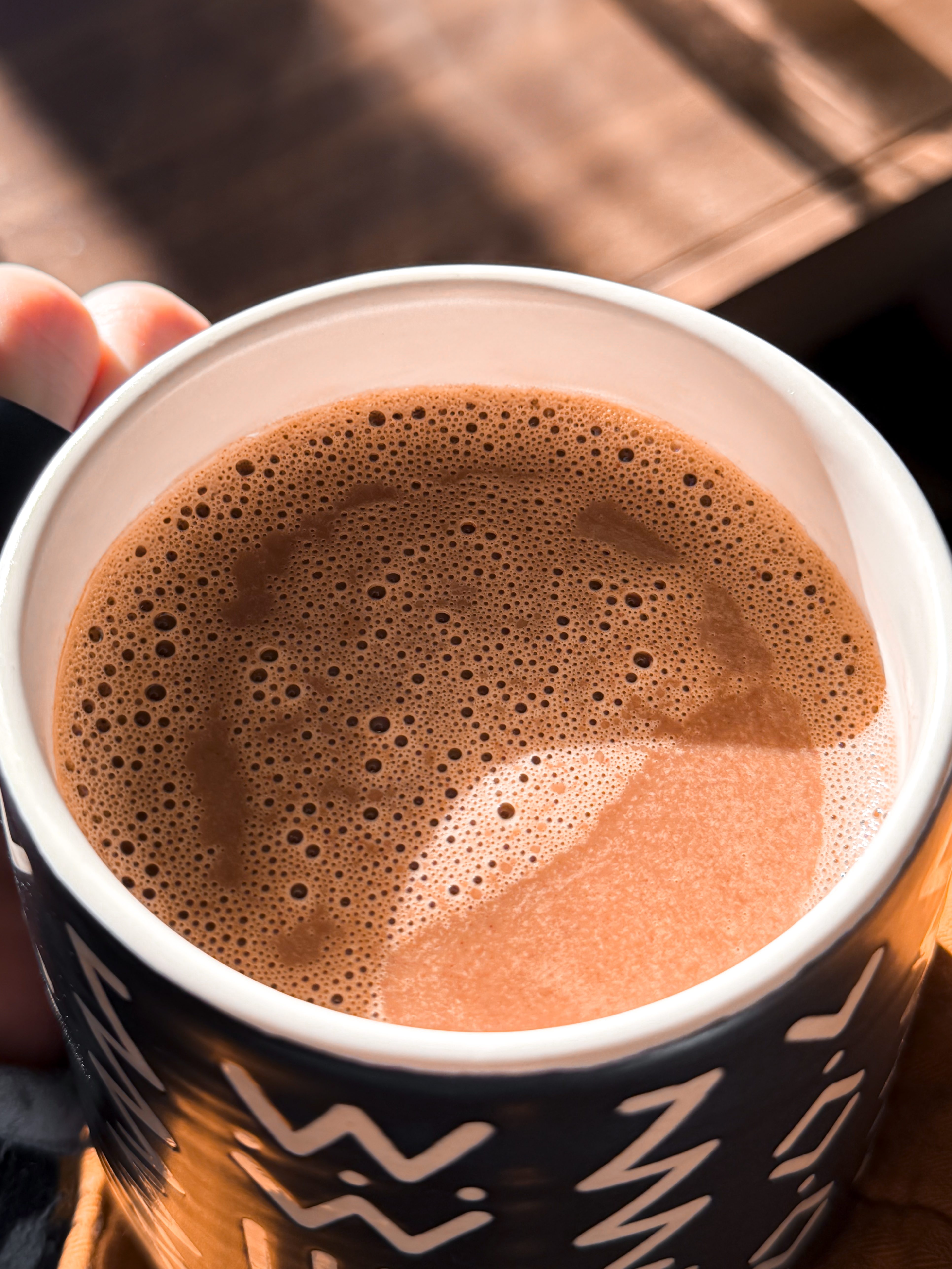 A close-up of frothy hot chocolate in a black mug with white geometric patterns.